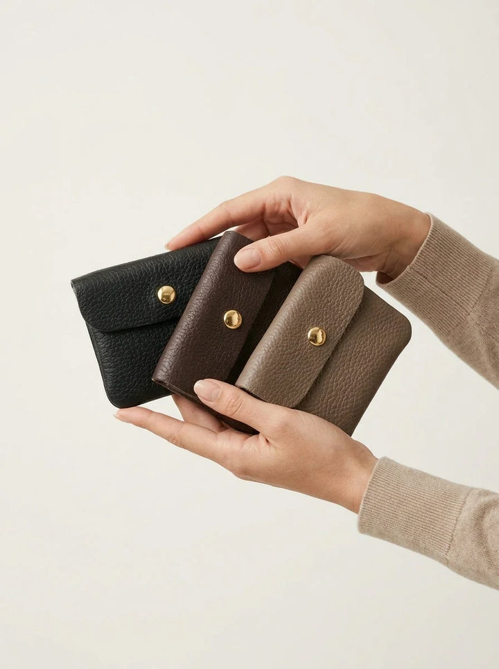 Mãos a segurar três carteiras em pele Noble Mini nas cores preto, castanho e bege